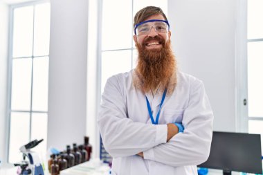 Young redhead man wearing scientist uniform standing with arms crossed gesture at laboratory