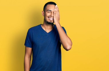 Young african american man wearing casual t shirt covering one eye with hand, confident smile on face and surprise emotion. 