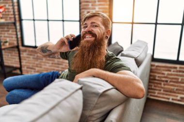 Genç kızıl saçlı, gülümseyen, kendinden emin bir adam evde akıllı telefondan konuşuyor.