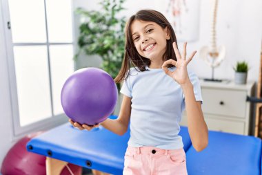 Young hispanic girl holding small rehabilitation ball doing ok sign with fingers, smiling friendly gesturing excellent symbol 