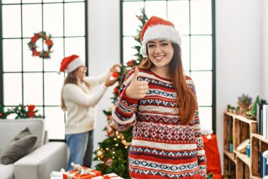 Beautiful couple at home standing by christmas tree smiling happy and positive, thumb up doing excellent and approval sign 