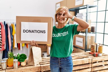 Middle age blonde woman wearing volunteer t shirt holding donations box smiling happy doing ok sign with hand on eye looking through fingers 