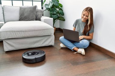Evde dizüstü bilgisayarla elektrikli süpürgeyi kontrol eden beyaz bir kız oturuyor..