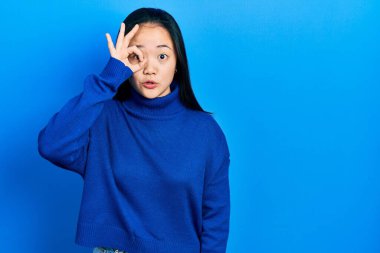 Young chinese girl wearing casual clothes doing ok gesture shocked with surprised face, eye looking through fingers. unbelieving expression. 