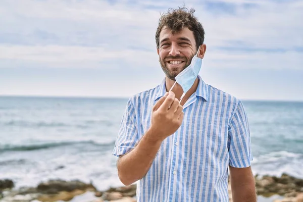 Jovem Hispânico Sorrindo Feliz Segurando Máscara Médica Praia — Fotografia de Stock