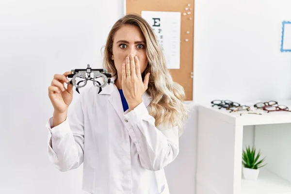 Young Beautiful Optician Woman Holding Optometry Glasses Covering Mouth Hand — стоковое фото