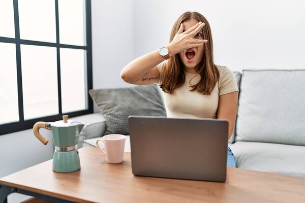 Молодая брюнетка, сидящая дома с ноутбуком, пьет чашку кофе, подглядывая рукой в лицо и глаза, смотрящие сквозь пальцы со смущенным выражением лица.. 