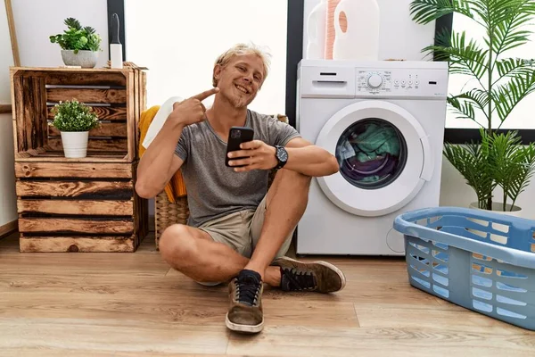 Young Blond Man Doing Laundry Using Smartphone Smiling Cheerful Showing — 스톡 사진
