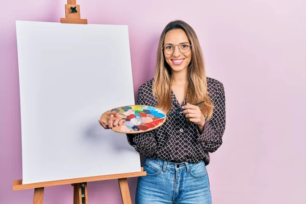 画家のイーゼルによるパレットで絵を描く美しいヒスパニック系の女性は 顔に幸せとクールな笑顔で笑顔立っています 歯を見せて — ストック写真