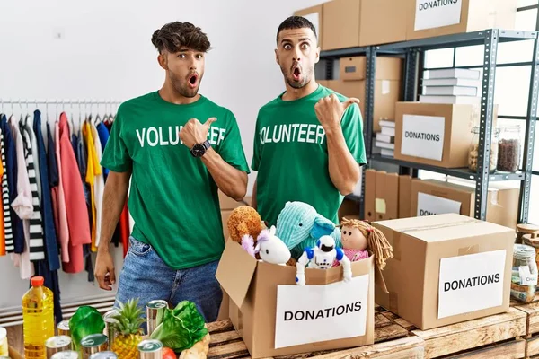 Young Gay Couple Wearing Volunteer Shirt Donations Stand Surprised Pointing — Zdjęcie stockowe