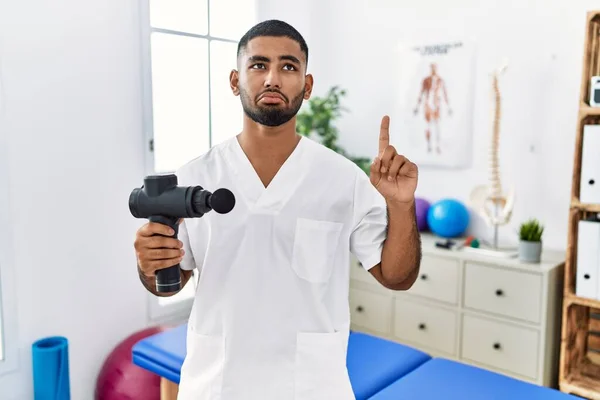 Young Indian Physiotherapist Holding Therapy Massage Gun Wellness Center Pointing — стоковое фото
