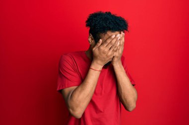 Young african american man with beard wearing casual red t shirt with sad expression covering face with hands while crying. depression concept. 