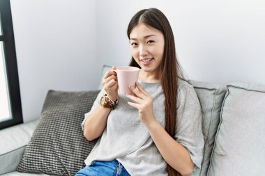 Genç Asyalı kadın oturma odasındaki kanepede kahve içiyor.