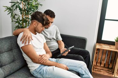 Two hispanic men couple using touchpad hugging each other at home