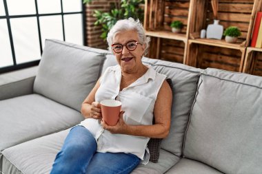 Yaşlı gri saçlı kadın evdeki kanepede kahve içiyor.