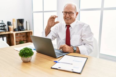 Senior man working at the office using computer laptop smiling and confident gesturing with hand doing small size sign with fingers looking and the camera. measure concept. 