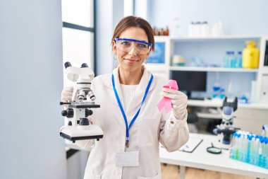 Young brunette woman working at scientist laboratory holding pink ribbon relaxed with serious expression on face. simple and natural looking at the camera. 
