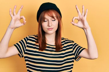 Kızıl saçlı genç kadın yün şapka takıyor rahatlıyor ve gözleri kapalı gülümsüyor parmaklarıyla meditasyon hareketi yapıyor. yoga konsepti. 