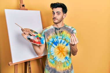 Young hispanic man standing drawing with palette by painter easel stand doing money gesture with hands, asking for salary payment, millionaire business 