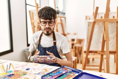 Hispanic man with beard at art studio with hand on stomach because indigestion, painful illness feeling unwell. ache concept. 