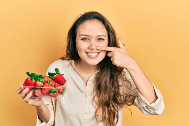 Young hispanic girl holding strawberries pointing with hand finger to face and nose, smiling cheerful. beauty concept 