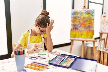 Young brunette teenager at art studio surprised with hand on head for mistake, remember error. forgot, bad memory concept. 