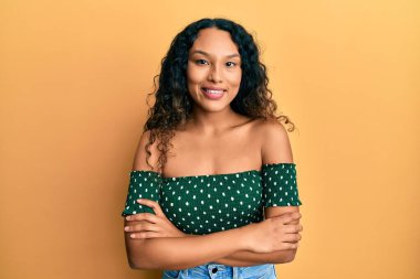 Gündelik kıyafetler giyen, kollarını kavuşturup kameraya gülümseyen genç Latin kadın. pozitif kişi. 