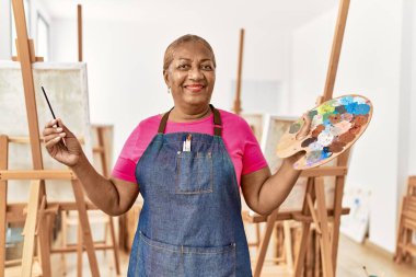 Kıdemli Afro-Amerikan kadın sanat stüdyosunda palet ve boya fırçası tutarak gülümsüyor.