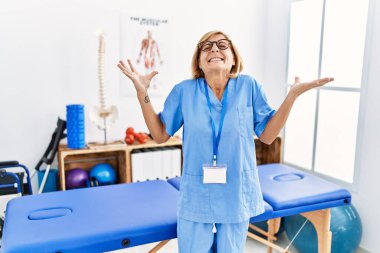 Middle age blonde woman working at pain recovery clinic celebrating mad and crazy for success with arms raised and closed eyes screaming excited. winner concept 
