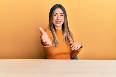 Young hispanic woman wearing casual clothes sitting on the table smiling cheerful offering palm hand giving assistance and acceptance. 