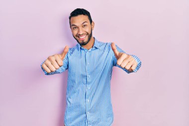 Hispanic man with beard wearing casual blue shirt approving doing positive gesture with hand, thumbs up smiling and happy for success. winner gesture. 