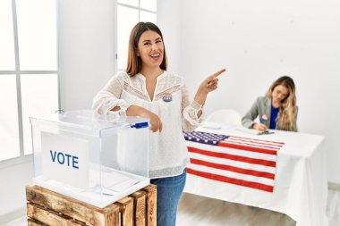 Young brunette woman voting putting envelop in ballot box smiling happy pointing with hand and finger to the side 