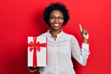Young african american woman holding gift smiling with an idea or question pointing finger with happy face, number one 