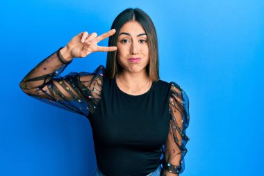 Young hispanic woman with victory gesture fingers on eye puffing cheeks with funny face. mouth inflated with air, catching air. 