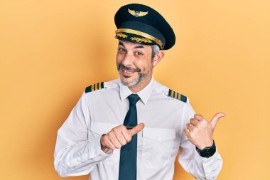 Handsome middle age man with grey hair wearing airplane pilot uniform pointing to the back behind with hand and thumbs up, smiling confident 