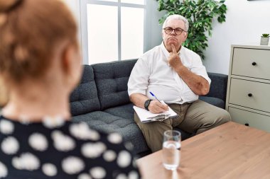 Senior psychologist man at consultation office thinking concentrated about doubt with finger on chin and looking up wondering 