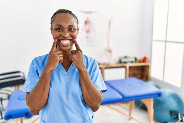 Black woman with braids working at pain recovery clinic smiling with open mouth, fingers pointing and forcing cheerful smile 