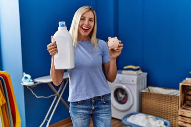 Beautiful woman doing laundry holding detergent bottle and piggy bank smiling and laughing hard out loud because funny crazy joke. 