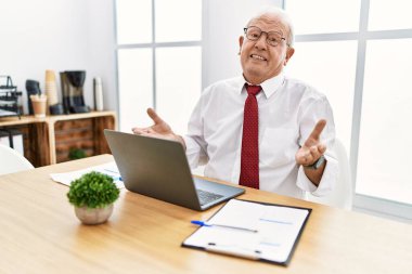 Senior man working at the office using computer laptop smiling cheerful with open arms as friendly welcome, positive and confident greetings 