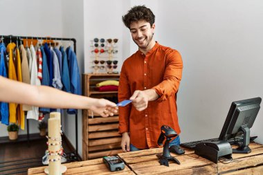 Young hispanic shopkeeper man giving credit card to customer at clothing store.