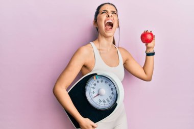 Young hispanic woman wearing sportswear holding weighing machine and apple angry and mad screaming frustrated and furious, shouting with anger looking up. 
