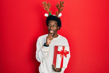 Young african american man wearing deer christmas hat holding gift with hand on chin thinking about question, pensive expression. smiling and thoughtful face. doubt concept. 
