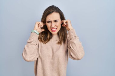 Young woman standing over isolated background covering ears with fingers with annoyed expression for the noise of loud music. deaf concept. 