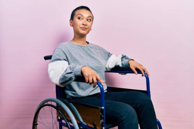 Beautiful hispanic woman with short hair sitting on wheelchair smiling looking to the side and staring away thinking. 