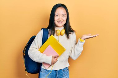 Young chinese girl holding student backpack and books smiling cheerful presenting and pointing with palm of hand looking at the camera. 