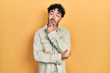 Elleri çenesinde gündelik giysiler içinde soru soran genç İspanyol bir adam, düşünceli bir ifade. Düşünceli bir yüzle gülümsüyor. şüphe kavramı. 