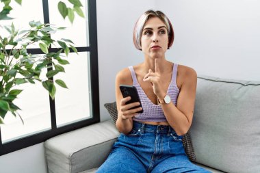 Young beautiful woman using smartphone typing message sitting on the sofa thinking concentrated about doubt with finger on chin and looking up wondering 