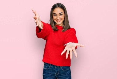 Beautiful brunette woman wearing casual winter sweater looking at the camera smiling with open arms for hug. cheerful expression embracing happiness. 