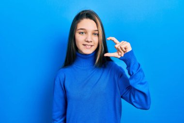 Young brunette girl wearing turtleneck sweater smiling and confident gesturing with hand doing small size sign with fingers looking and the camera. measure concept. 