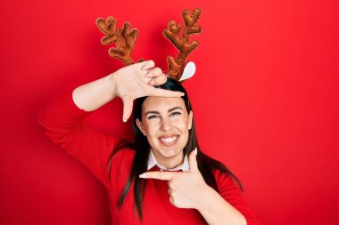 Young hispanic woman wearing cute christmas reindeer horns smiling making frame with hands and fingers with happy face. creativity and photography concept. 